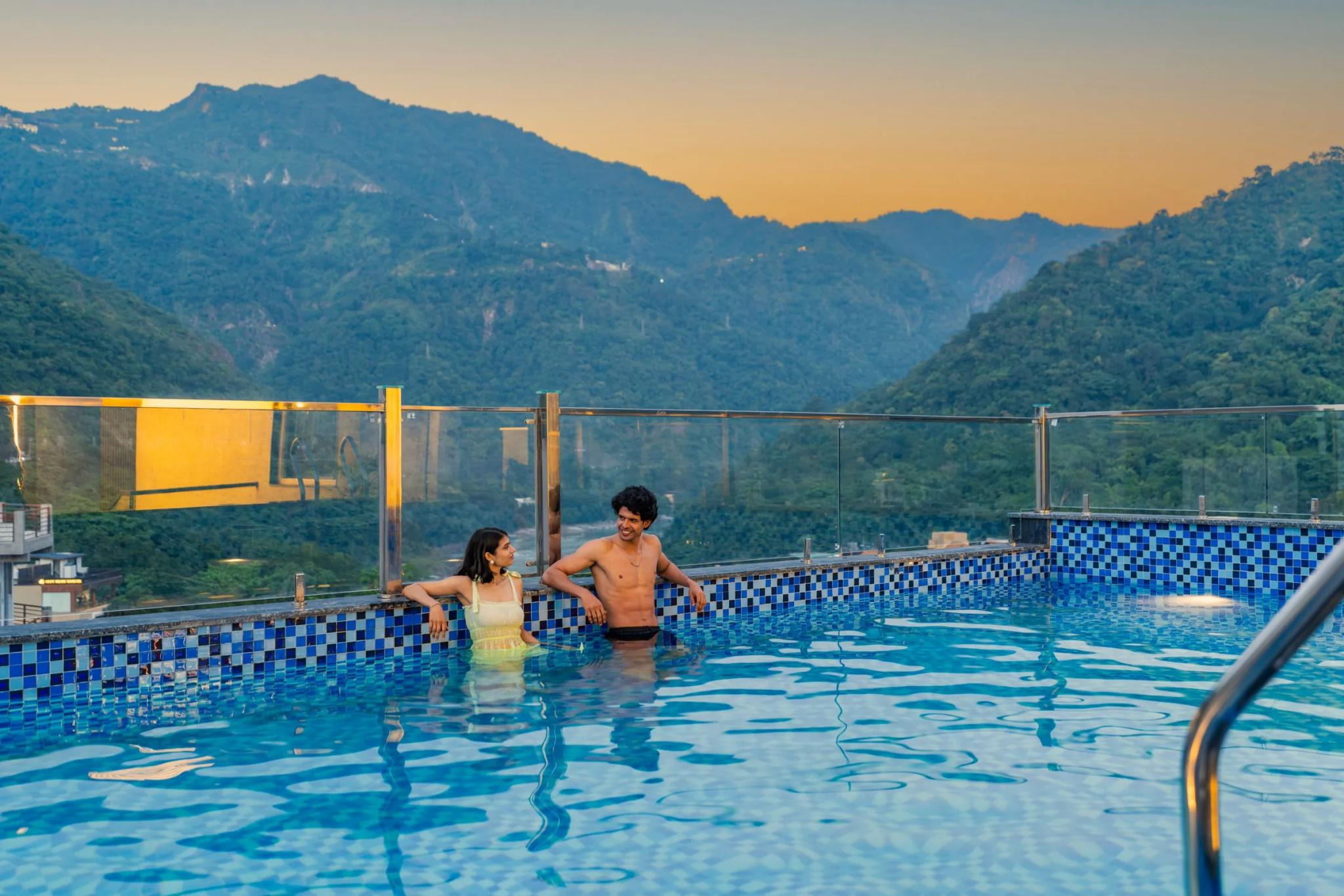 Two friends chatting in the rooftop pool at golden hour, mountains behind